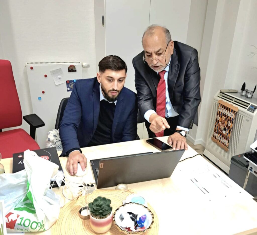 Two men in an office looking at a laptop on a cluttered wooden desk, one pointing at the screen while the other sits nearby.