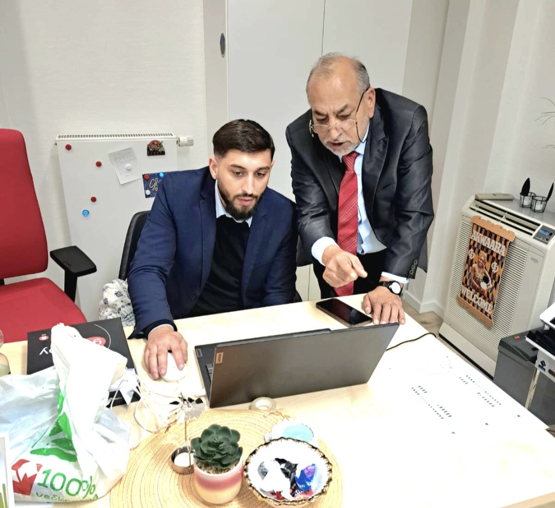Two men in an office looking at a laptop on a cluttered wooden desk, one pointing at the screen while the other sits nearby.