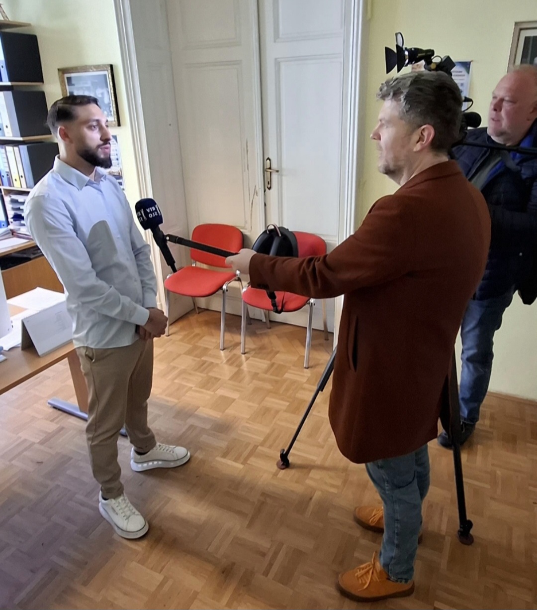 Man in a light blue shirt answers questions as a reporter holds a handheld microphone in an office setting; a cameraman films in the background with a tripod.