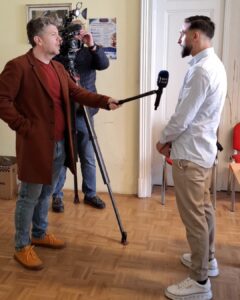 Reporter interviewing a man indoors with a handheld microphone labeled RTV SLO, camera crew filming in the background.