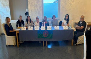 Panel discussion with eight people seated at a head table, name placards, flags, and a circular emblem on a blue banner.