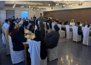 People seated around long, U-shaped tables listening to a presenter at a podium in a conference room with exposed ceiling lights and string bulbs.